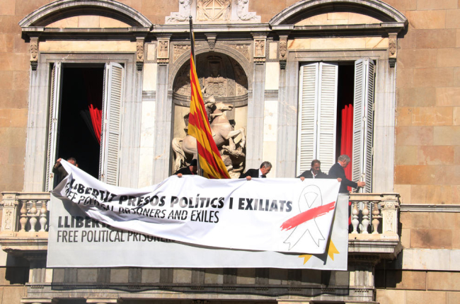 Trabajadores del Gobierno añaden una pancarta encima del anterior, ahora con el lazo blanco y una franja roja, en el balcón del Palau de la Generalitat.
