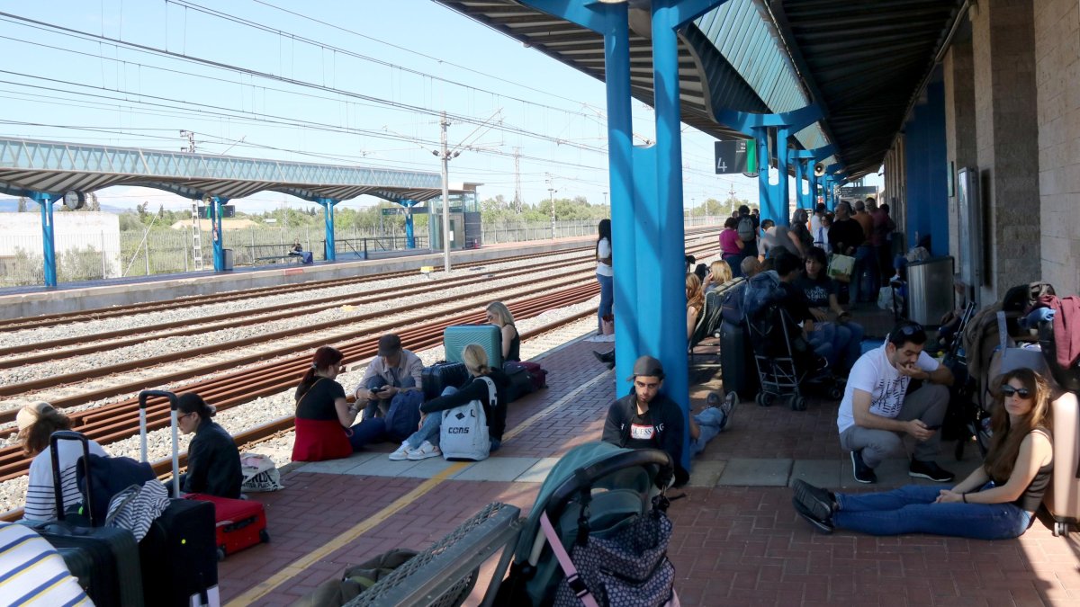 Viajeros sentados en el andén de la estación de l'Aldea después del apagón eléctrico y el paro de sus trenes este lunes al mediodía