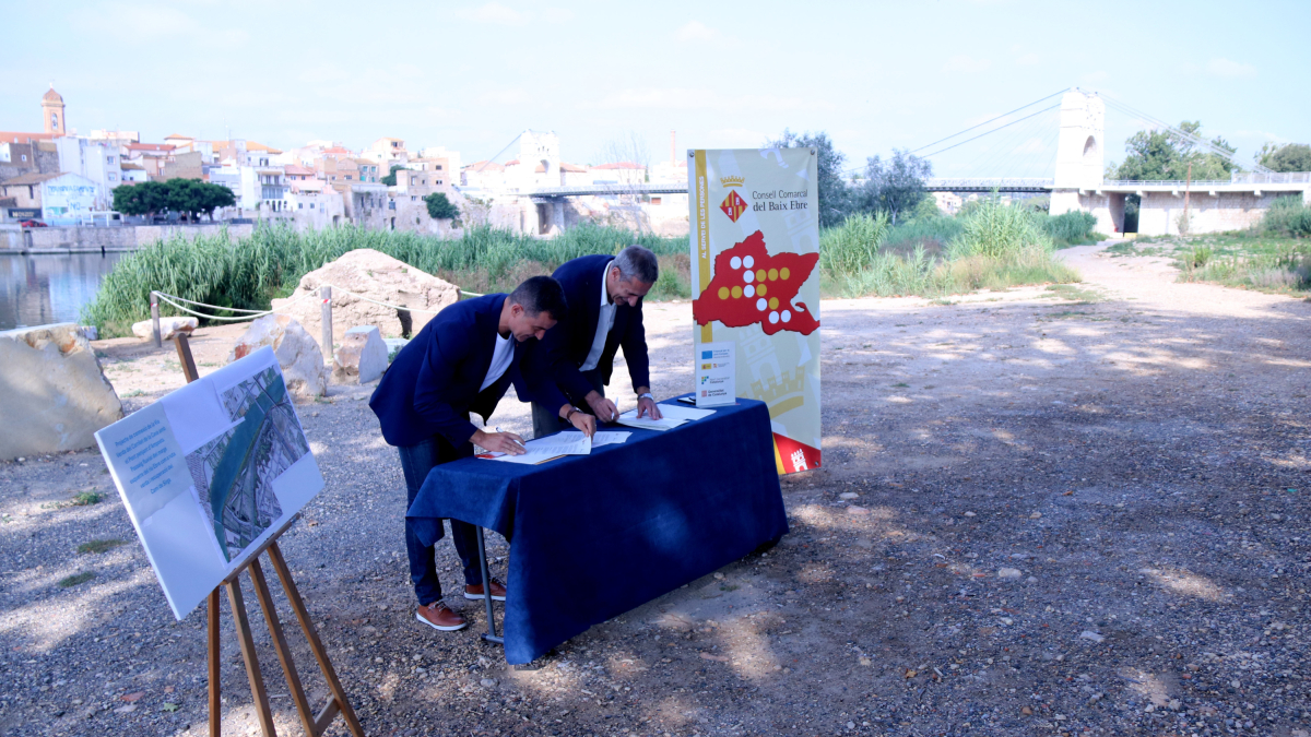 El presidente del Consejo del Baix Ebre, Antoni Gilabert, y el alcalde de l'Aldea, Xavier Royo, firman la cesión de los terrenos con el puente colgante de Amposta y el tramo del enlace de la vía verde, en el fondo.