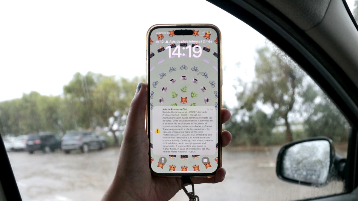 Un teléfono móvil, con la alerta de Protección Civil, y una calle llena de agua de lluvia en el fondo, en Amposta