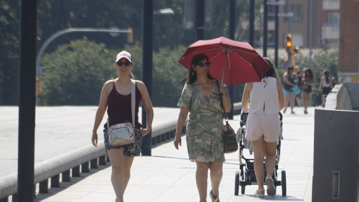 Una mujer se protege del sol con un paraguas y otra con una gorra, mientras caminan por el puente Viejo de Lleida.