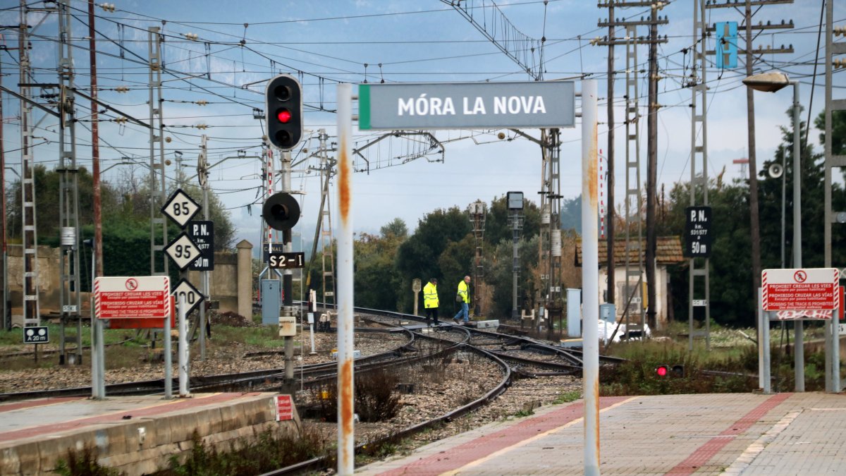 Carteles en la estación de tren de la R-15 en Móra la Nova, con dos operarios en el fondo haciendo comprobaciones en las vías