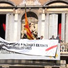 Treballadors del Govern afegeixen una pancarta damunt l'anterior, ara amb el llaç blanc i una franja vermella, al balcó del Palau de la Generalitat.
